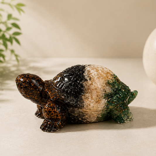 A small decorative turtle figurine with a glossy, marbled shell in black, beige, and green, placed on a light beige surface with a blurred plant and ceramic pot in the background.